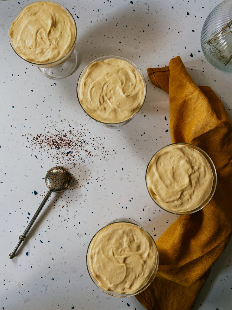 tiramisu in glasses with a yellow napkin and a chocolate powder dispenser, a hand reaching for it with a spoon