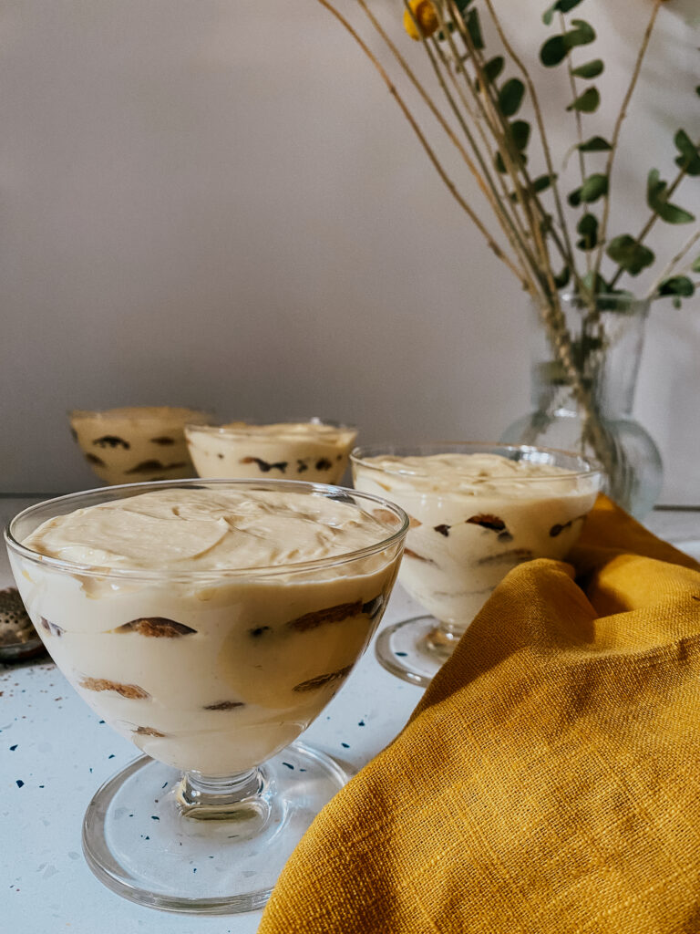 tiramisu in glasses with a yellow napkin and a chocolate powder dispenser, a vase with dried flowers in the background