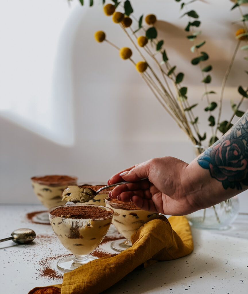 tiramisu in glasses with a yellow napkin and a chocolate powder dispenser, a hand reaching for it with a spoon