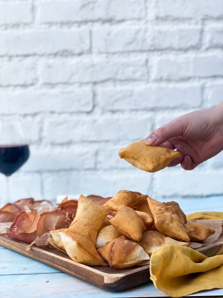 a hand holding a gnocco fritto with prosciutto in it, on the background a glass of red wine and under a board of meat and cheese