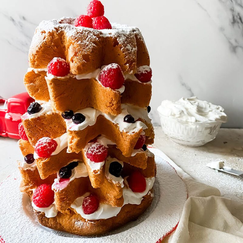 pandoro farcito on a plate, under a towel, in the back a spatula with cream and a bowl with whipped cream, on a marble background
