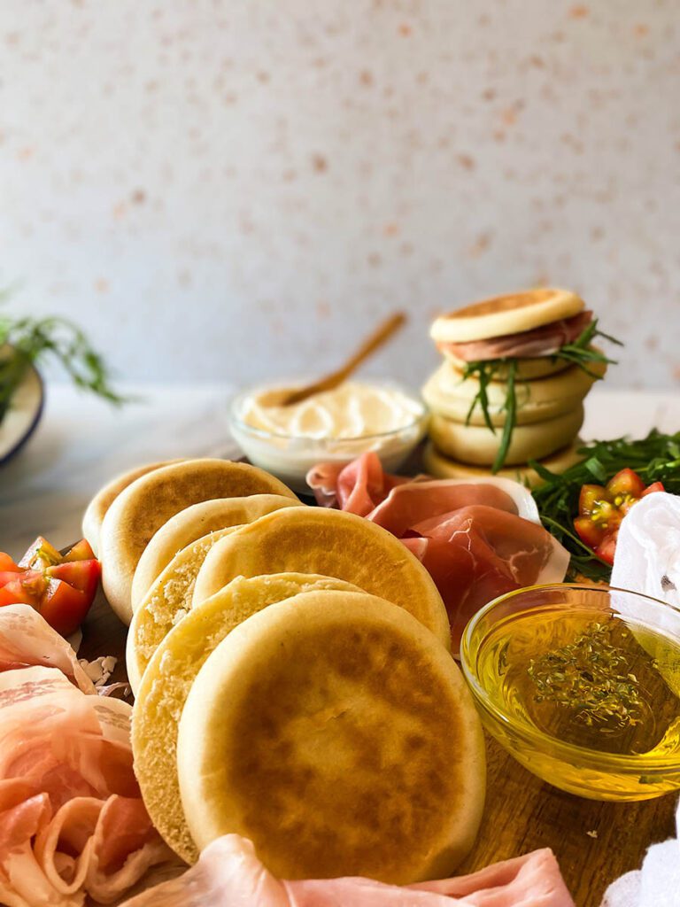 a wooden board filled with freshly made tigelle and all the bits and trims to fill them with including cured meats, cheese and olive oil