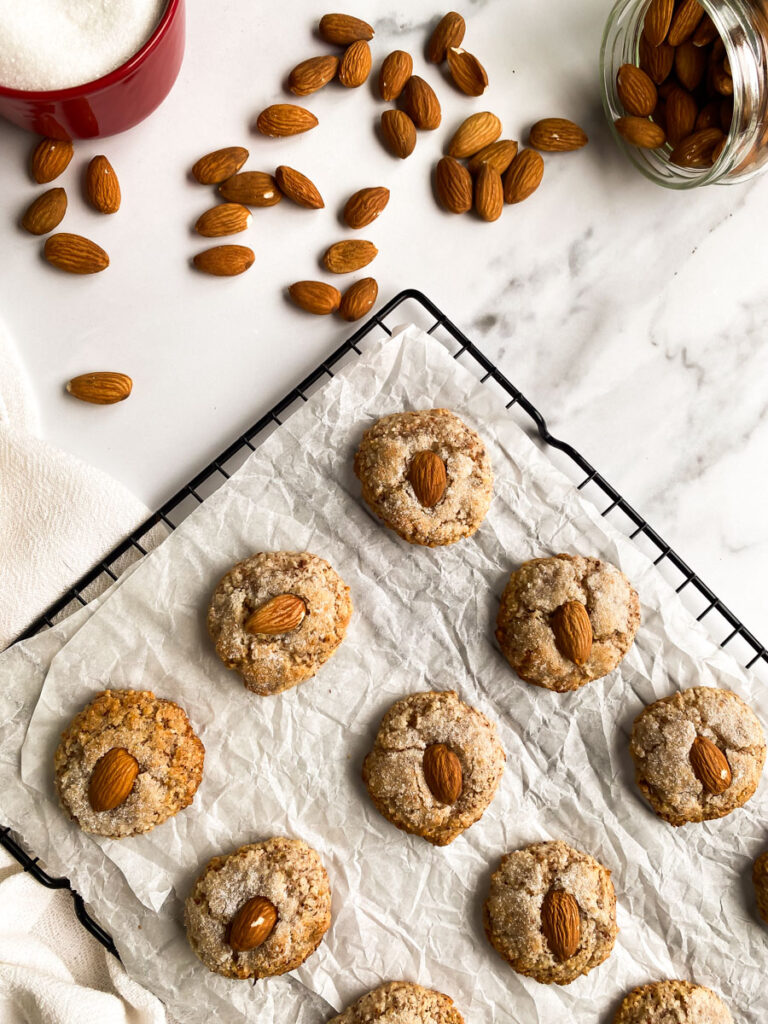 from the top amaretti sardi on parchment paper, laying on a grid, a white towel around, more almonds and sugar in pots