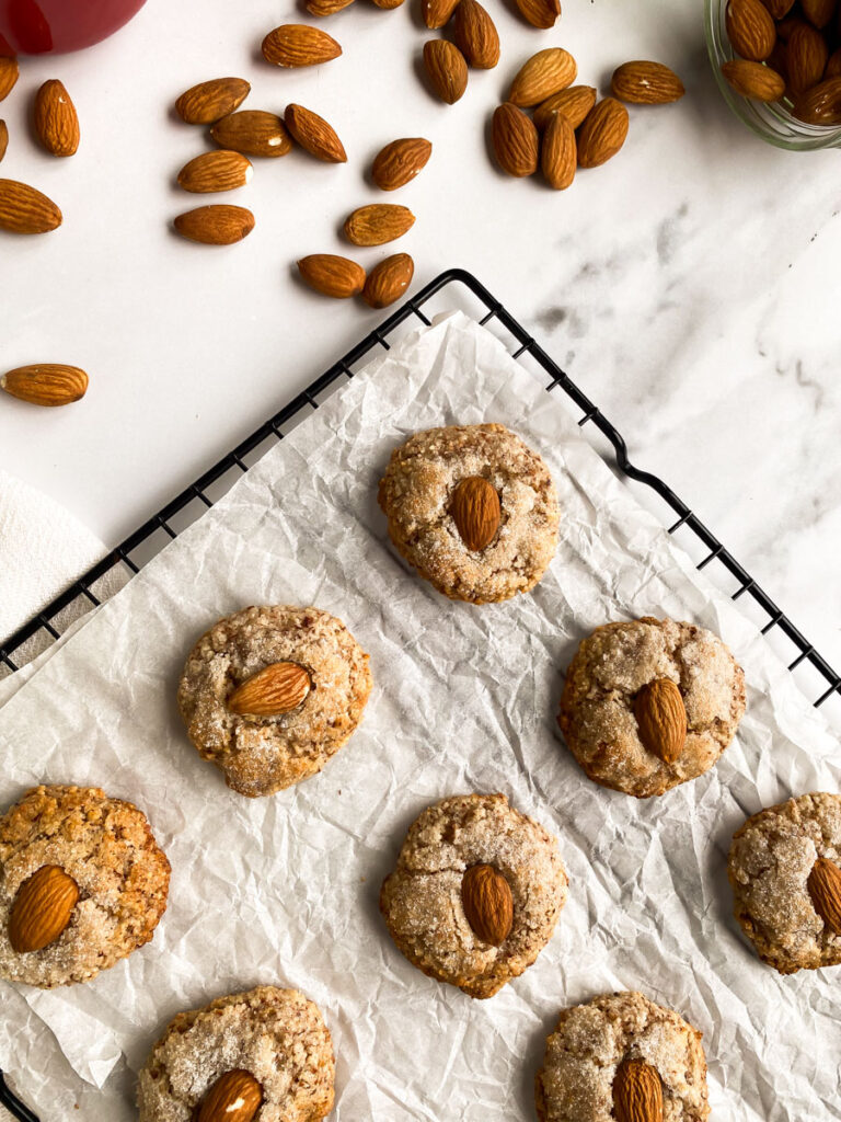 above, amaretti sardi on parchment paper, laying on a grid, a white towel around, more almonds and sugar in pots