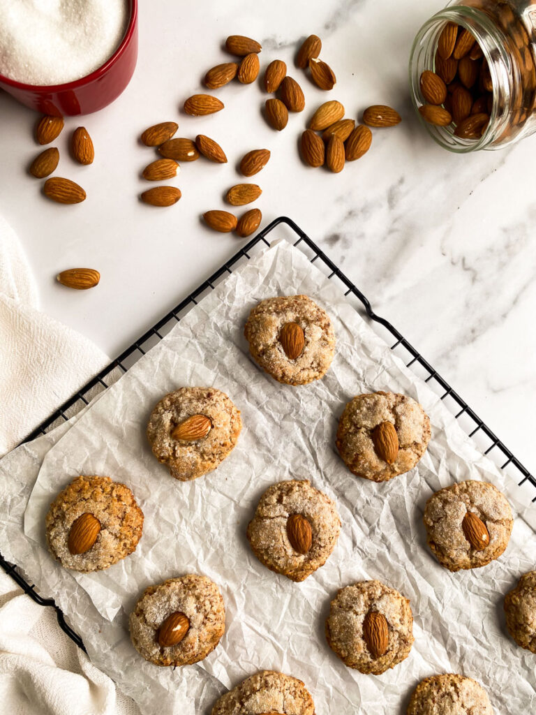amaretti sardi on parchment paper, laying on a grid, a white towel around, more almonds and sugar in pots
