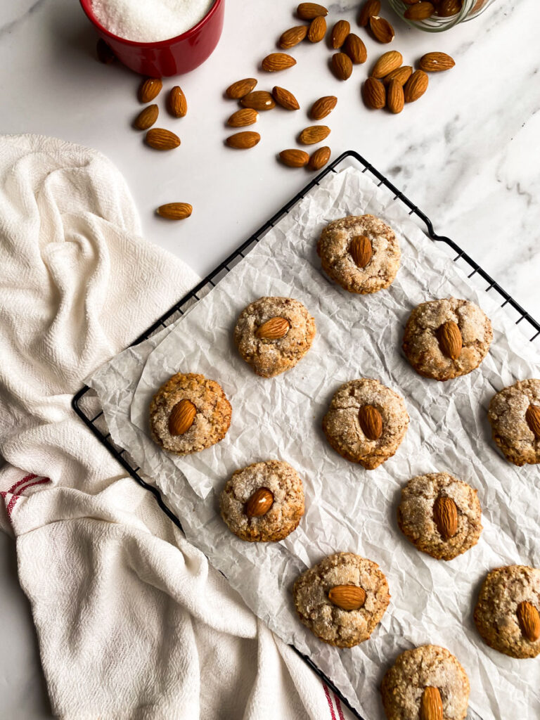amaretti sardi on parchment paper, laying on a grid, a white towel around, more almonds and sugar in pots