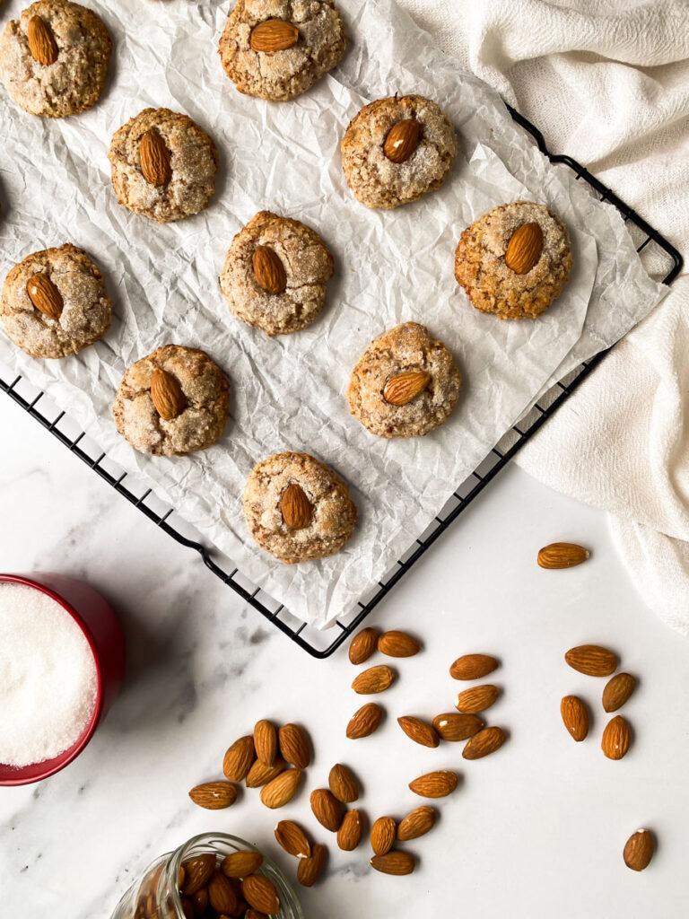 above, amaretti sardi on parchment paper, laying on a grid, a white towel around, more almonds and sugar in pots
