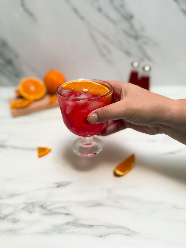 Hand holding a Blood orange spritz in two glasses, blood oranges behind and two bottles of bitter