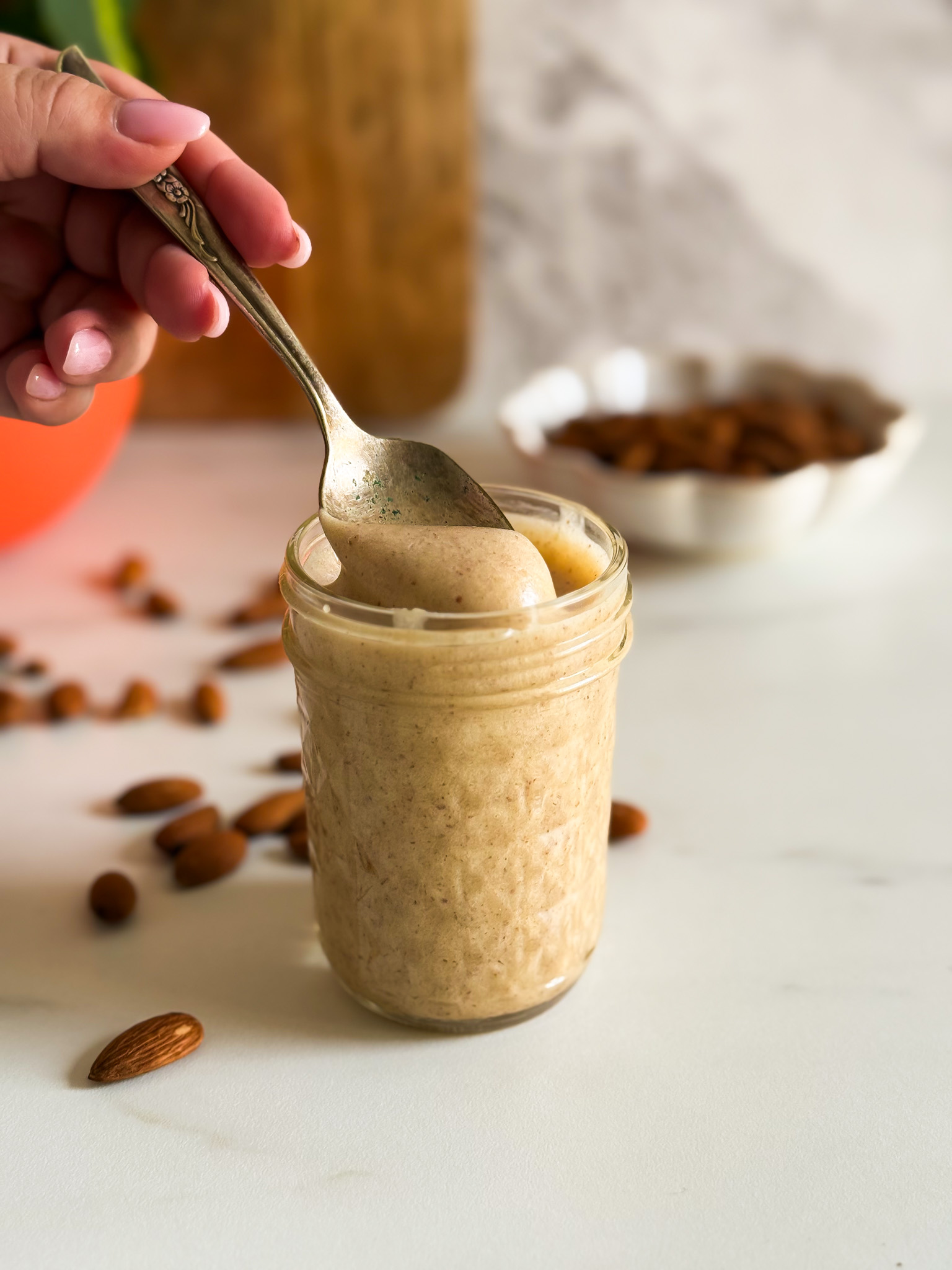 close up detail almond cream in a jar, hand holding a spoon and lifting some of the cream, almonds, a bowl of almonds, plant and wooden board on the background