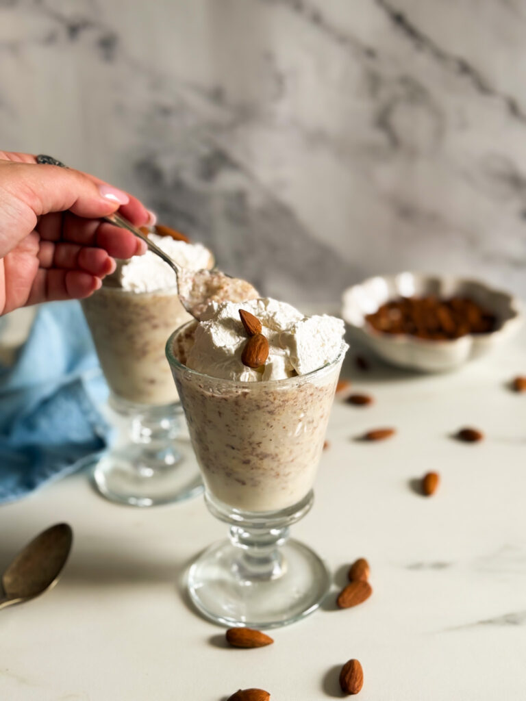 hand holdinga. spoonful of granita, granita di mandorle in two tall glasses, spoon, almonds and cream in the background