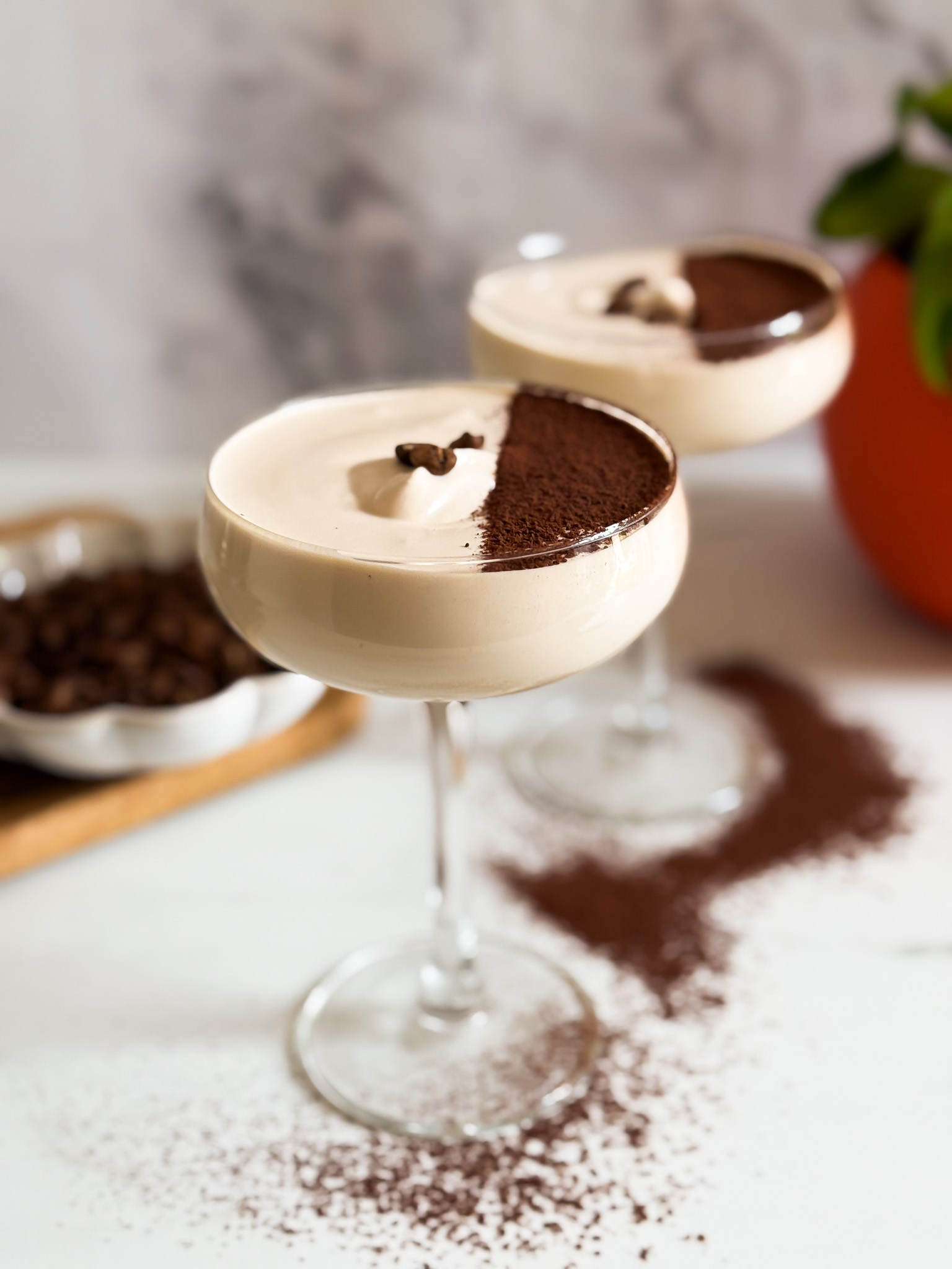front view two coupes of crema al caffe hand with a spoon reaching for some, coffee beans in the background and chocolate powder around