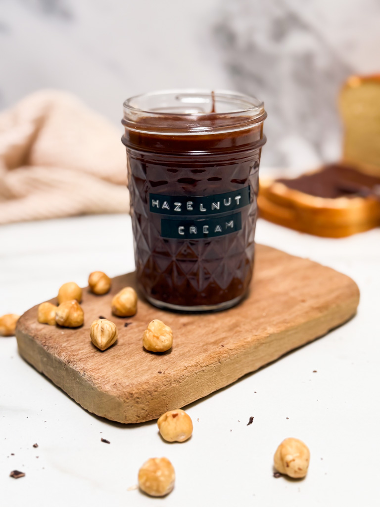 close up of a jar of hazelnut cream on a wooden board, hazelnuts scattered around, bread with hazelnut cream on the background with a towel