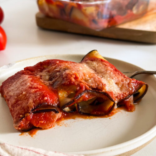 close up plate of involtini melanzane with a fork on the side, towel, a tray with more eggplant rollups behind and tomatoes