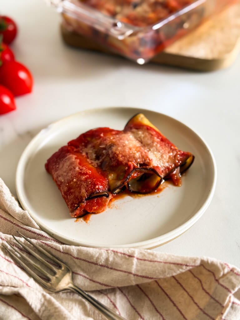 plate of involtini melanzane with a fork on the towel, a tray with more eggplant rollups behind and tomatoes