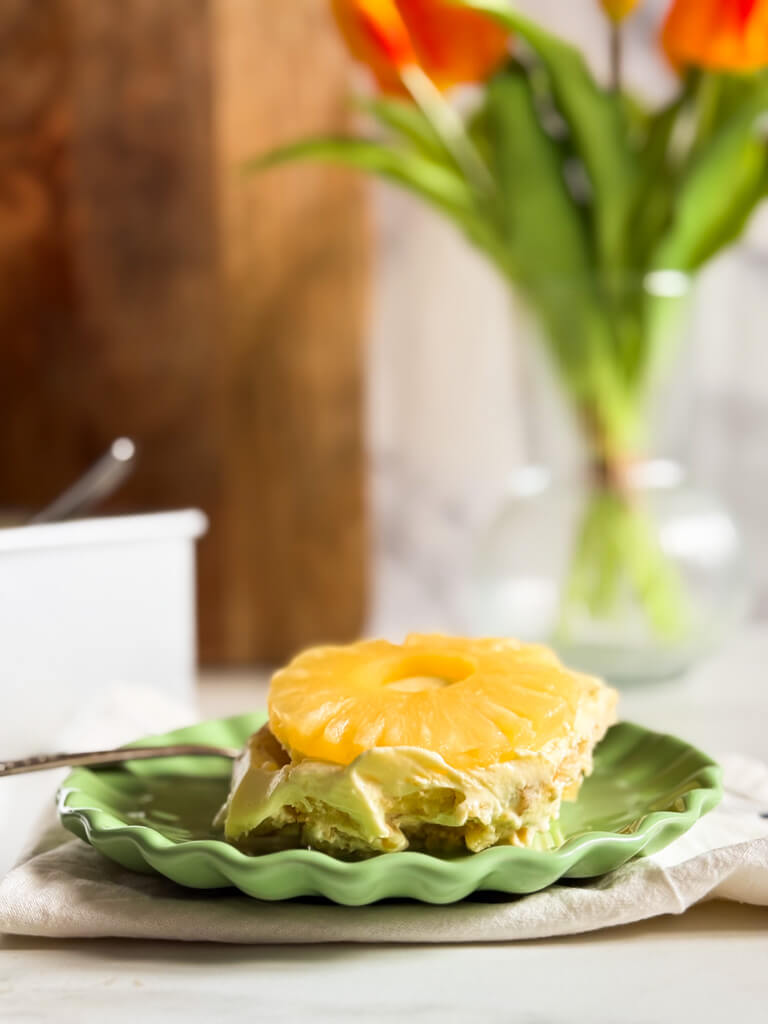 from front - pineapple tiramisu on a green plate, on top of a towel with a spoon behind, in the background the tiramisu in a tray, wooden board and a vase with tulips