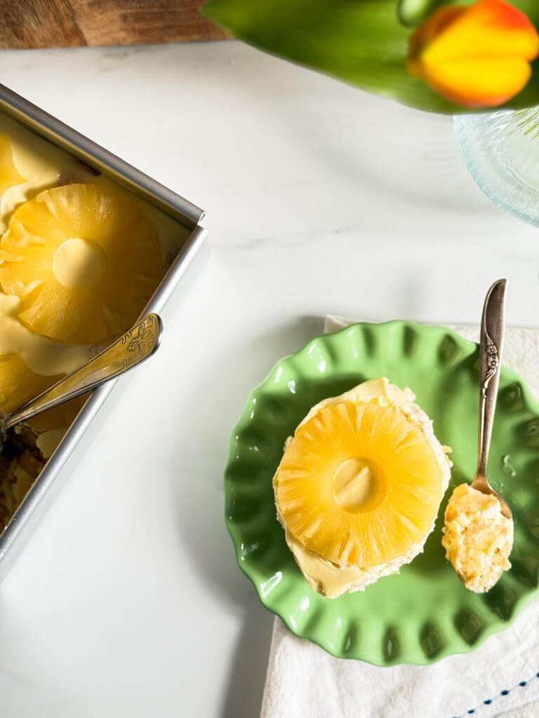 from top - pineapple tiramisu on a green plate, on top of a towel with a spoon beside it with a piece, in the background the tiramisu in a tray, wooden board and a vase with tulips