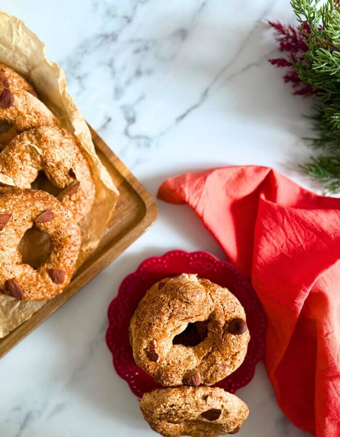 from top stack of roccocò cookies on a red plate and a red towel, in the background more cookies on a wooden board and a christmas tree ornament, on a marble background