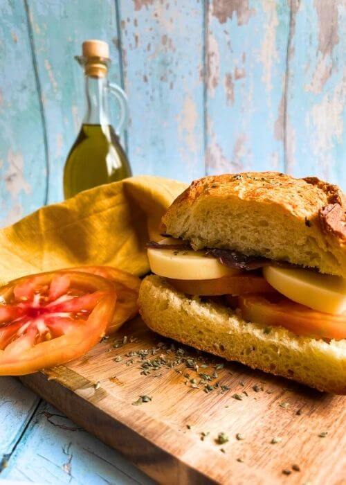 pane cunzato on a wooden board, tomatoes, yellow towel and bottle of olive oil