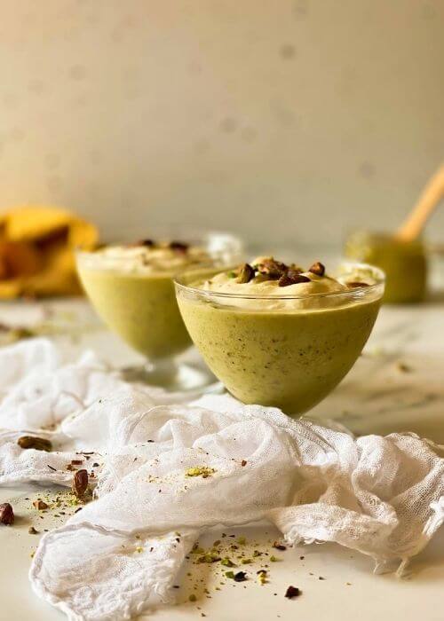 two glasses of pistachio granita, towels and ground pistachios around, at the back a jar of pistachio cream
