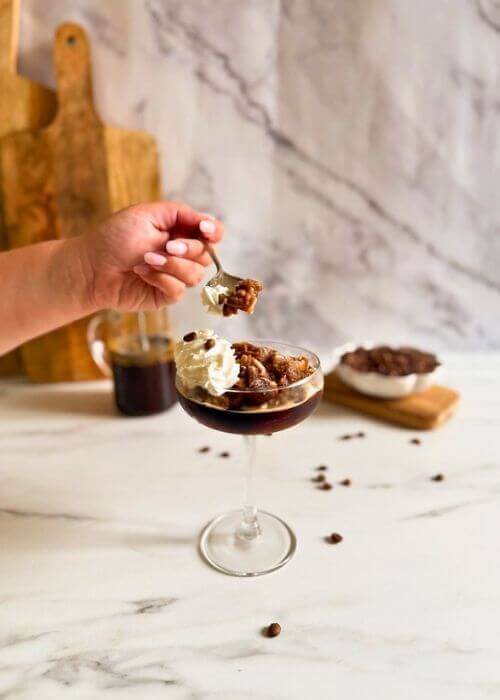 coupe of coffee granita granita al caffè with whipped cream, hand with a spoon holding up some, coffee beans in a pot and coffee pot in the background