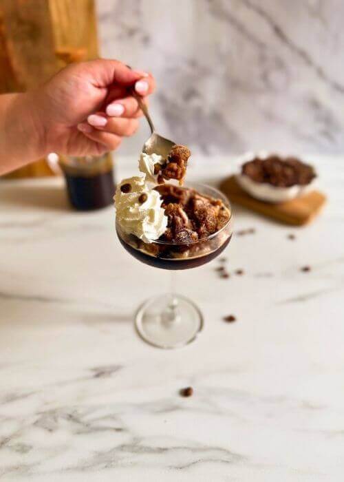 coupe of coffee granita granita al caffè with whipped cream, hand with a spoon holding some, coffee beans in a pot and coffee pot in the background