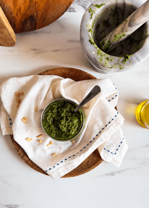 from top low glass container with fresh homemade pesto alla genovese, pine nuts and towel around, wooden board, olive oil and mortar and pestel in the background