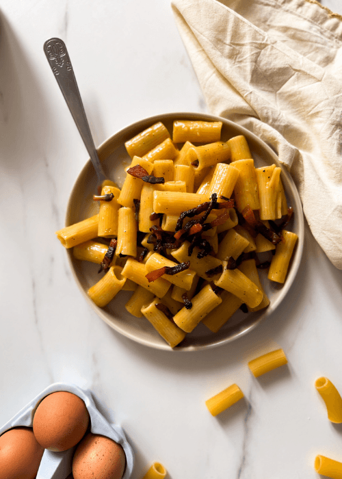 from top view rigatoni alla carbonara on a plate with a fork, towel, eggs and dried pasta on the background