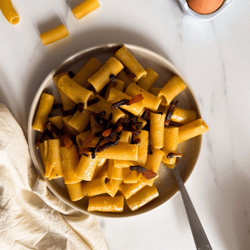 rigatoni alla carbonara on a plate with a fork, towel, eggs and dried pasta on the background