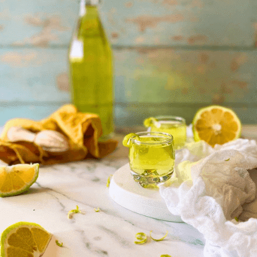 two glasses of limoncello on a marble plate, white and yellow towels around, lemon squeezer, lemons and lemon peel around, a bottle of limoncello in the background
