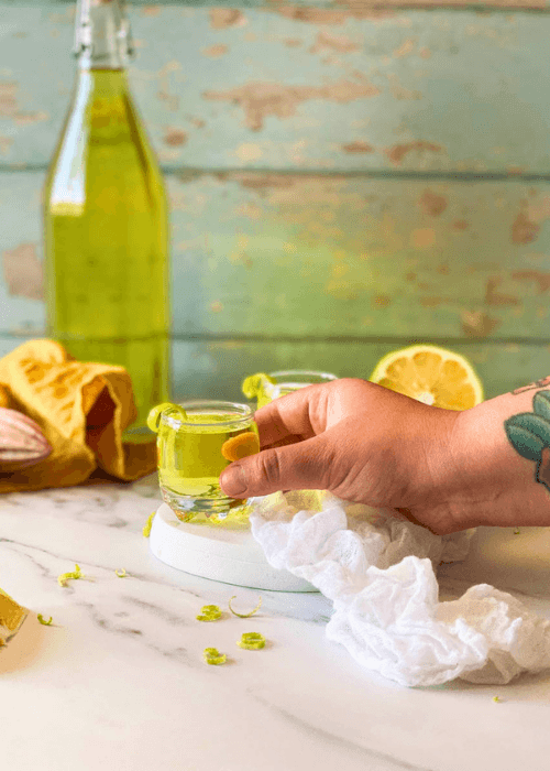 hand reaching for a glass, two glasses of limoncello on a marble plate, white and yellow towels around, lemon squeezer, lemons and lemon peel around, a bottle of limoncello in the background
