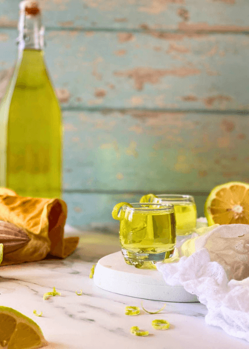 two glasses of limoncello on a marble plate, white and yellow towels around, lemon squeezer, lemons and lemon peel around, a bottle of limoncello in the background