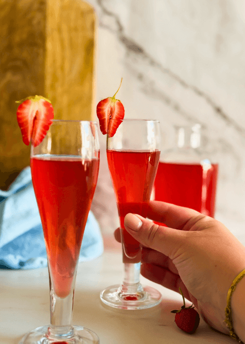 hand holding one strawberry liqueur in two flute glasses, strawberries to decor, a bottle with liqueur, wooden board and a towel on the background
