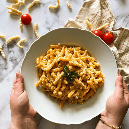 close up, hands holding the plate, plate of busiate pasta with red pesto, tomatoes and dried busiate around, towel under