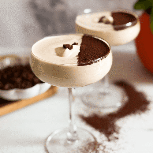 front view two coupes of crema al caffe hand with a spoon reaching for some, coffee beans in the background and chocolate powder around
