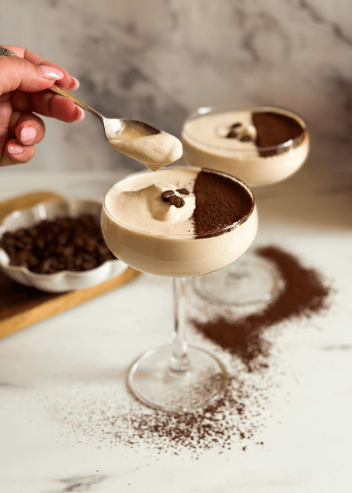 two coupes of crema al caffe hand with a spoon reaching for some, coffee beans in the background and chocolate powder around