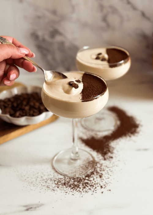 two coupes of crema al caffe hand with a spoon taking some, coffee beans in the background and chocolate powder around